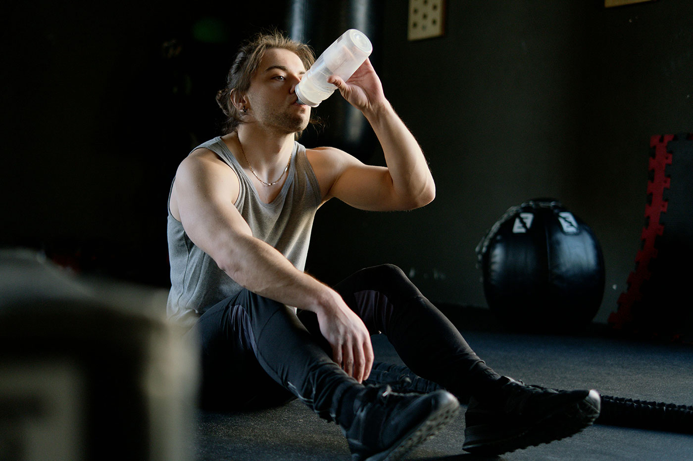 млад мъж седнал на земята във фитнес залата пие вода от бутилка
